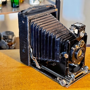 Op de afbeelding: Een vintage vouwcamera met een zwart met leer beklede behuizing en een zwarte accordeonbalg. De camera is voorzien van een ronde lens, metalen instelknoppen en een zoeker aan de bovenzijde, gemonteerd op een scharnierende basis.