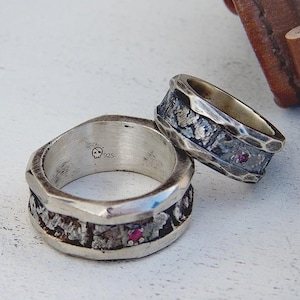 Op de afbeelding: Twee sterling zilveren ringen met een textuur bandontwerp. Eén ring heeft een kleine schedel en de stempel "925". De ringen hebben een gehamerde metalen textuur met ingebedde rode edelstenen. De ringen liggen op een wit oppervlak.