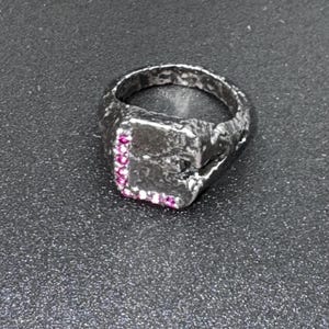 Könnte beinhalten: Ein silberfarbener Ring mit quadratischer Fassung, besetzt mit kleinen rosa Edelsteinen. Der Ring hat ein strukturiertes, gehämmertes Aussehen. Die Edelsteine sind in einer Reihe entlang einer Kante der quadratischen Fassung angeordnet. Der Ring ist ein Schmuckstück.