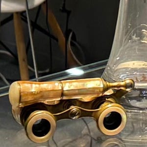 Puede incluir: Prismáticos de ópera antiguos de latón con un barril de nácar. Los prismáticos tienen un diseño detallado con dos lentes circulares y un mecanismo de enfoque central. El latón tiene una pátina cálida y envejecida.