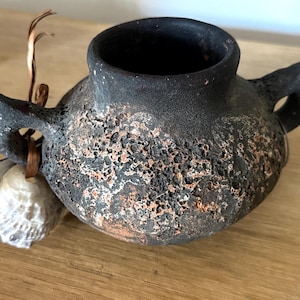 Może przedstawiać: Mały, okrągły, czarny ceramiczny garnek z dwoma uchwytami po bokach i szorstką, porowatą, teksturowaną powierzchnią z widocznymi odcieniami czerwonawego brązu. Biała muszla jest przywiązana do jednego uchwytu cienką brązową wstążką z rafii.