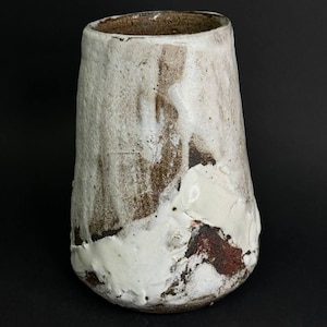 Pode incluir: Um vaso de cerâmica cilíndrico com uma superfície texturizada e irregular. O exterior apresenta uma base de argila castanha e escura parcialmente coberta por um esmalte branco espesso e escorrido que revela partes do material terroso.