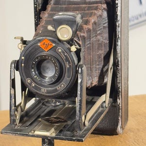 May include: A vintage Agfa folding camera with a black leather-covered body and a dark brown accordion-style bellows. The lens assembly features a black circular housing with the text Agfa, Jgestar, and various aperture settings clearly visible.