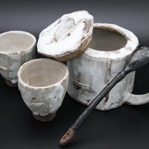 Peut inclure: Un service à thé en céramique fait main comprenant deux petites tasses, un pot avec couvercle et anse, et une longue cuillère sombre et texturée. Les pièces présentent un émail blanc rustique et irrégulier avec des tons d'argile brune.
