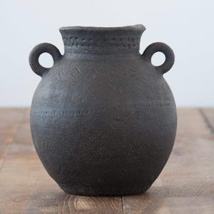 Peut inclure: Un vase en céramique de couleur charbon foncé avec deux anses en forme de boucle. Le vase a un corps arrondi et une surface texturée avec des détails gravés autour du col et de la section médiane. Le vase repose sur une surface en bois.