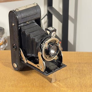 May include: A vintage folding camera with a black leather-textured body and a black bellows mechanism. The front features a circular lens assembly with metallic silver and black detailing, including a small viewfinder and mechanical adjustment dials.