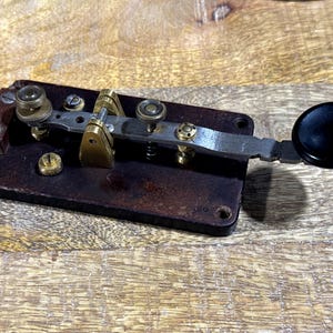 Puede incluir: Llave telegráfica antigua con base de madera marrón oscuro y componentes metálicos. La llave presenta detalles de latón, una palanca plateada y un pomo negro. Diseñada para enviar código Morse.