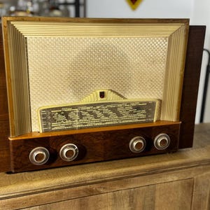 Könnte beinhalten: Ein Vintage-Tischradio mit einem dunklen Holzgehäuse, einem cremefarbenen Lautsprechergitter mit horizontalen Rillen in der Mitte und einer horizontalen Glasskala. Vier runde Drehknöpfe befinden sich an der unteren vorderen Holzblende.
