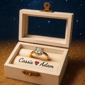 May include: A gold engagement ring with a large, clear gemstone sits in a white wooden box. The box is open, revealing the ring on a cream-colored cushion. The box is inscribed with the names "Cassie ❤️ Adam."