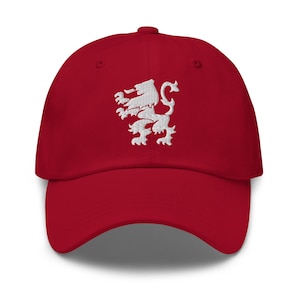 May include: A deep red baseball cap featuring a white embroidered lion design centered on the front panel. The cap has a curved brim, a structured crown with ventilation eyelets, and a button at the top, showcasing a classic six-panel construction.