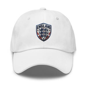 Pode incluir: Boné de beisebol branco com o brasão da seleção inglesa. O brasão tem a forma de um escudo com a palavra "ENGLAND" no topo e inclui o emblema dos Três Leões e uma bola de futebol.