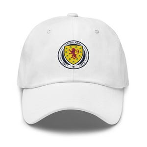 Könnte beinhalten: Eine weiße Baseballkappe mit einem kreisförmigen Emblem auf der Vorderseite. Das Emblem zeigt einen gelben Schild mit einem roten, aufsteigenden Löwen, umgeben von einem dunkelblauen Rand mit dem Text SCOTLAND und EST 1873 in weißen Buchstaben.