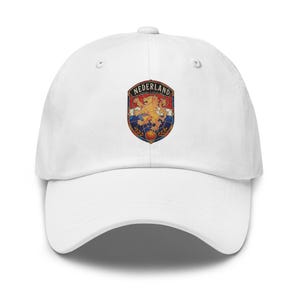 May include: A white baseball cap featuring a centered crest graphic. The crest displays a golden lion on a red, white, and blue background with the text NEDERLAND arched at the top. The design has a stained glass aesthetic with dark outlines.