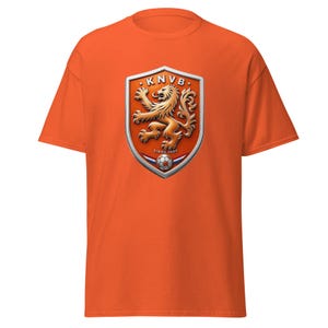Könnte beinhalten: Orangefarbenes T-Shirt mit dem KNVB-Logo, einem Schild mit einem Löwen und einem Fußball. Das Logo ist in Orange-, Gold- und Silbertönen gehalten. Der Text "SINDS 1889" befindet sich unter dem Fußball. Das Shirt ist einfarbig orange.