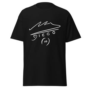 Puede incluir: Camiseta negra con un gráfico de firma blanca, incluyendo el nombre "DIEGO" y el número "10". El diseño está centrado. La camiseta está hecha de una tela suave.