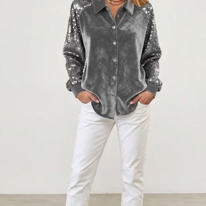 Puede incluir: Una camisa gris de terciopelo con mangas de lentejuelas se combina con pantalones blancos. La camisa tiene cuello y está abotonada. Las mangas están adornadas con lentejuelas plateadas. Los pantalones son rectos y blancos.