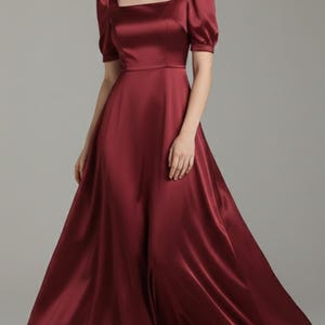 May include: A full-length, burgundy satin dress featuring a square neckline and short, puffed sleeves. The dress has a fitted bodice and a flowing skirt. The model is wearing beige heels. Suitable for formal occasions.
