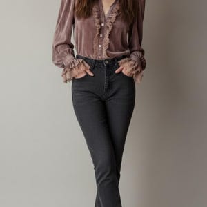 Può includere: Una donna indossa una camicetta di velluto malva con finiture in pizzo e jeans grigio scuro. La camicetta ha una chiusura con bottoni e maniche lunghe. I jeans sono a gamba dritta. Indossa anche stivaletti grigi.