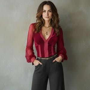 Könnte beinhalten: Eine bordeauxfarbene Samtbluse mit transparenten langen Ärmeln und einem V-Ausschnitt. Die Bluse hat ein figurbetontes Oberteil mit vertikalen Einsätzen und eine zarte Spitzenborte. Das Model trägt eine schwarze Hose und einen goldenen Gürtel.