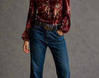Pode incluir: Uma blusa de veludo bordô com estampa floral, combinada com jeans azuis e um cinto decorativo. A blusa de manga comprida tem decote em V e está por dentro dos jeans. Os jeans têm perna larga. A modelo usa um colar.