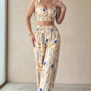 May include: A cream-coloured two-piece floral outfit, comprising a crop top with tie straps and high-waisted trousers. The set is decorated with blue, orange, and yellow floral patterns. The trousers have a wide-leg design.