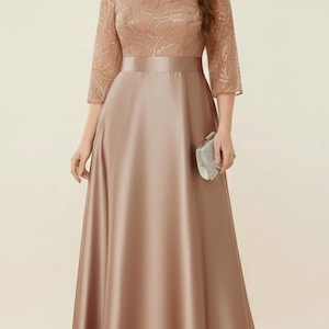 May include: A full-length, rose-toned evening gown. The bodice has a detailed, textured overlay with three-quarter sleeves. A satin ribbon defines the waist, and the skirt flows to the floor. A small, silver clutch bag completes the ensemble.