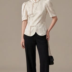 Könnte beinhalten: Cremefarbene Bluse mit kurzen Ärmeln, Stehkragen und dekorativen Froschverschlüssen, kombiniert mit schwarzer Hose mit weitem Bein. Die Bluse hat ein dezentes Blumenmuster. Eine kleine schwarze Handtasche wird getragen.