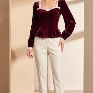 May include: A burgundy velvet top with long sleeves and a square neckline, trimmed with white lace. The top has a fitted bodice and a peplum waist. Paired with cream-coloured trousers and black heels, creating a sophisticated ensemble.