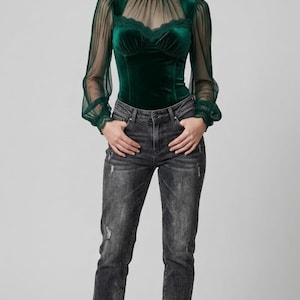 May include: A woman wearing a dark green velvet top with sheer sleeves and a high neckline, paired with distressed black jeans and black ankle boots. The top features lace detailing and a sweetheart neckline. The outfit is a mix of textures and styles.