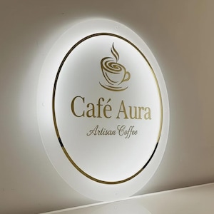 Può includere: Un'insegna rotonda e illuminata per "Café Aura Artisan Coffee". L'insegna presenta una grafica di tazza di caffè dorata e testo dorato. L'insegna è bianca con un bordo dorato ed è montata su un muro.