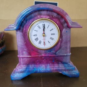 May include: A mantel clock with a swirling purple, pink, and blue resin frame featuring a gold-rimmed circular face. The white clock face displays black Roman numerals, black hour and minute hands, a red second hand, and the word QUARTZ.