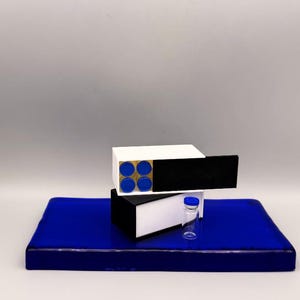 May include: A still-life composition featuring a deep blue rectangular base, upon which rests a white and black box with four blue circles. A small glass vial with a blue cap sits nearby. The background is a neutral gray.