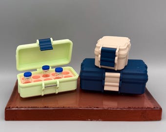 May include: A collection of small, colorful storage boxes. One is open, revealing vials inside. The boxes are in shades of green, blue, and beige, with secure latches. They sit on a reddish-brown rectangular base.