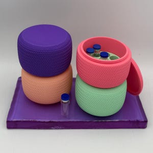 May include: A vibrant still life featuring a purple tray holding stacked, textured, circular containers in purple, peach, pink, and green. The pink container is open, revealing small vials. A single vial sits on the tray.