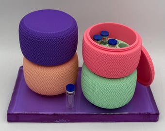 May include: A vibrant still life featuring a purple tray holding stacked, textured, circular containers in purple, peach, pink, and green. The pink container is open, revealing small vials. A single vial sits on the tray.