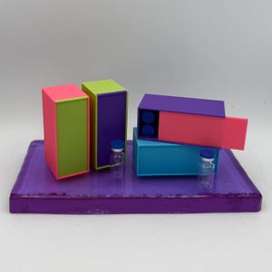 May include: A vibrant still life featuring colorful rectangular boxes in pink, green, purple, and blue, arranged on a purple tray. Two small glass vials with blue caps are also present. The boxes have contrasting color schemes.