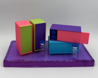 May include: A vibrant still life featuring colorful rectangular boxes in pink, green, purple, and blue, arranged on a purple tray. Two small glass vials with blue caps are also present. The boxes have contrasting color schemes.
