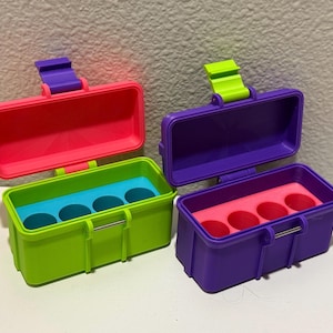 May include: Four colorful, rectangular storage containers with hinged lids. The containers are in shades of pink, green, and purple, with contrasting lid and interior colors. Each container has four circular compartments inside.