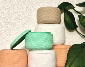 May include: A collection of small, round, lidded containers in various pastel colors: peach, mint green, white, and tan. Each container has a textured, woven-like pattern. One lid is open, revealing the inside of the container.