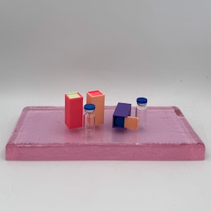 May include: A pink rectangular tray holds several small objects. There are two clear glass vials with blue caps, and three colorful rectangular boxes. The boxes are red, orange, and purple, with green, red, and orange accents. The tray has a textured surface.