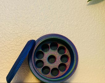 May include: A circular, multi-colored herb grinder with a gradient finish shifting from blue to green and purple. The open top reveals a central plate featuring eight outer circular holes and one larger central hole, with a textured chevron pattern.