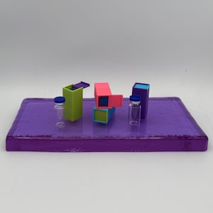 May include: A vibrant display featuring a rectangular purple tray with several colorful geometric shapes and two small glass vials with blue caps. The shapes are pink, green, blue, and purple. The vials are clear.