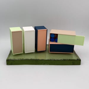 May include: A collection of colorful, geometric boxes in shades of green, white, navy, and peach, arranged on a green rectangular base. Some boxes are open, revealing blue circular objects inside. The boxes have a modern, minimalist design.