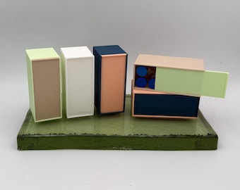 May include: A collection of colorful, geometric boxes in shades of green, white, navy, and peach, arranged on a green rectangular base. Some boxes are open, revealing blue circular objects inside. The boxes have a modern, minimalist design.