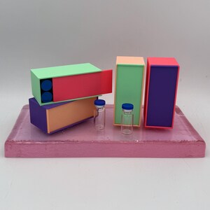May include: A still life composition featuring colorful, geometric boxes in green, red, purple, and peach, with two small glass vials with blue caps. The boxes are arranged on a pink rectangular base.