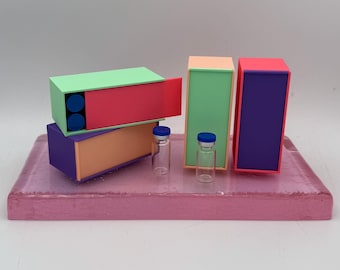 May include: A still life composition featuring colorful, geometric boxes in green, red, purple, and peach, with two small glass vials with blue caps. The boxes are arranged on a pink rectangular base.