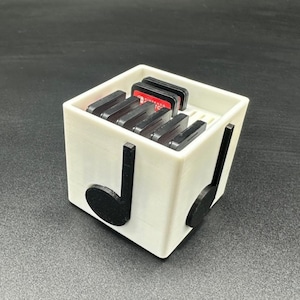 May include: A white, cube-shaped storage container with black musical note accents on the sides. The container holds several black and red game cartridges. The design is a creative blend of functionality and musical aesthetics.