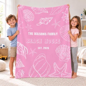 Peut inclure: Une couverture polaire rose ornée d'un motif en lignes blanches représentant divers coquillages et étoiles de mer. Le centre affiche le texte THE WILSONS FAMILY BEACH HOUSE EST. 2026 en lettres capitales blanches, créant un design côtier.