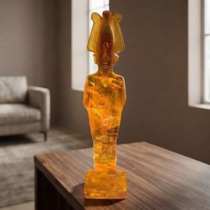 Puede incluir: Una figura translúcida de color ámbar que representa a un antiguo faraón egipcio con una corona alta y estilizada. La estatua presenta una superficie lisa y pulida con texturas internas y se apoya sobre una base rectangular.
