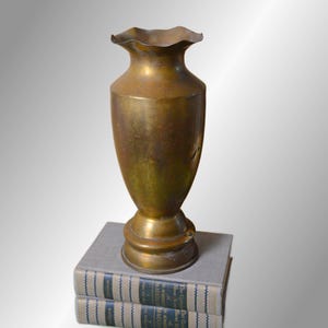 May include: A tall, metallic, brass-colored vase with a narrow neck and a wide, ruffled, scalloped rim. The vase features a smooth, tapered body that rests on a circular, pedestal-style base, showcasing a weathered, aged patina finish.